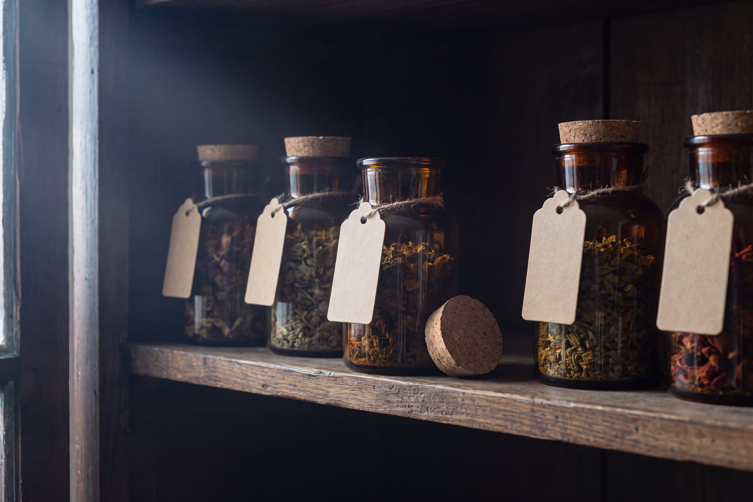 Cork-stoppered amber glass jars of dried herbs on a dark pantry shelf, stored away from light, heat and moisture.