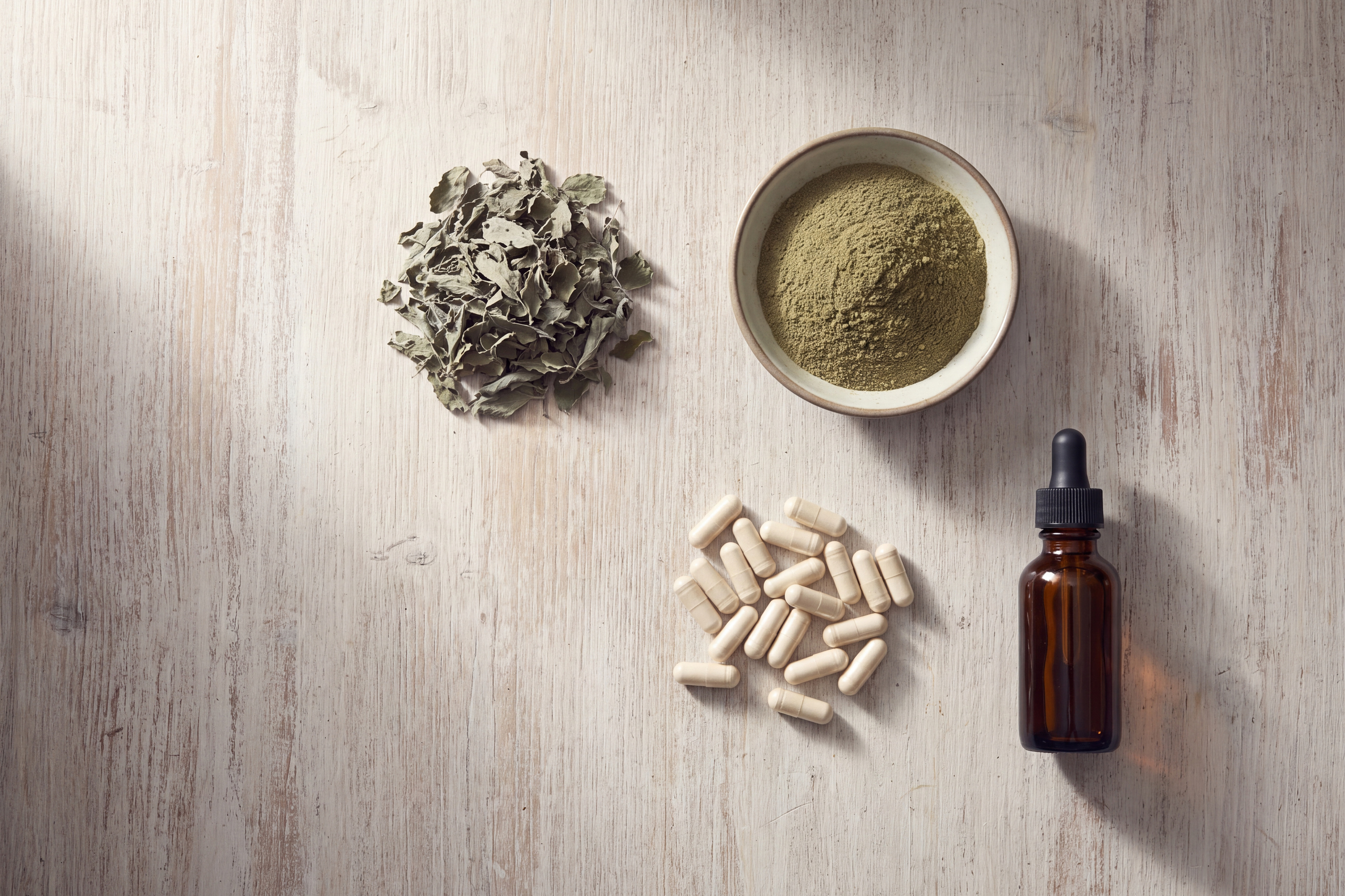 Dried holy tulsi leaf, milled powder, capsules and a tincture bottle arranged on a pale wooden surface.