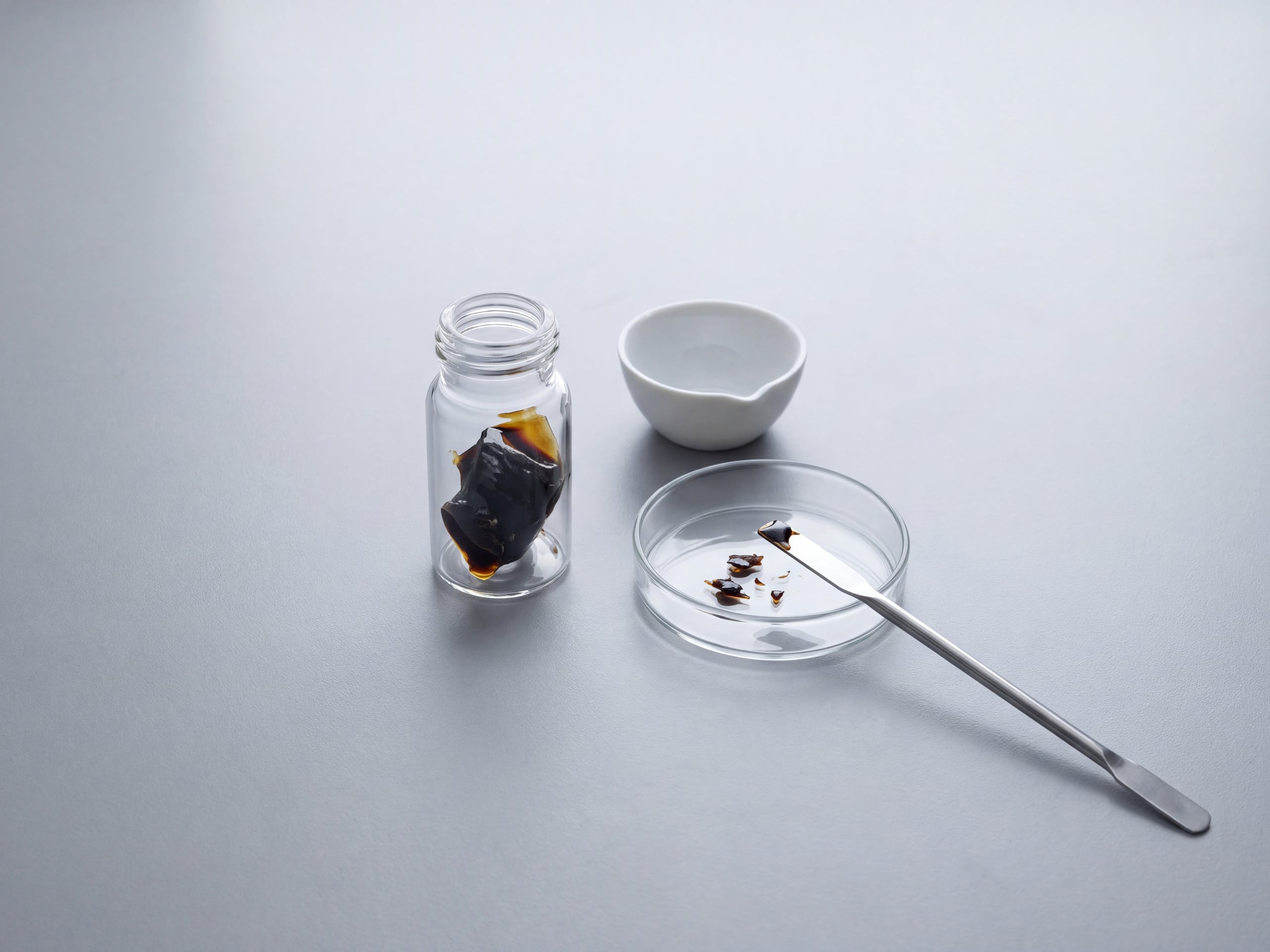 Dark shilajit resin in an unlabelled glass vial beside a porcelain crucible and spatula on a laboratory bench.
